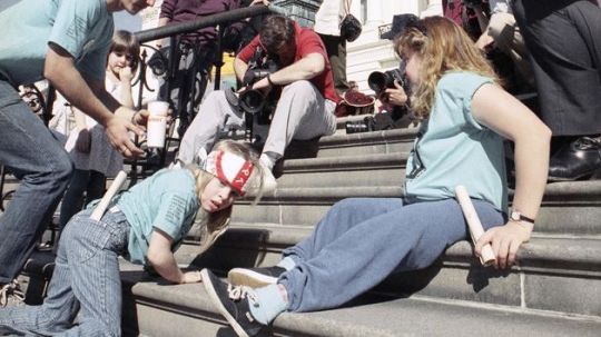  a small child with cerebral palsy is looking at the camera. She is wearing a light‐blue t‐shirt, jeans, and a bandanna. She is on her hands and knees as she crawls up the steps of the Capital Building as part of the Capital Crawl protest of 1990. 